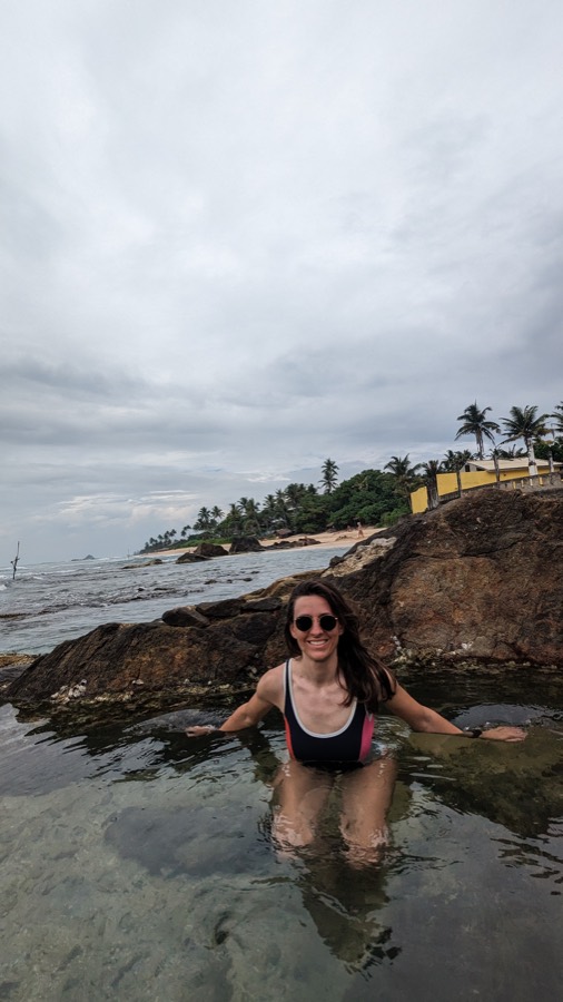 Alexandra sitting in a rock pool by a palm-fringed beach